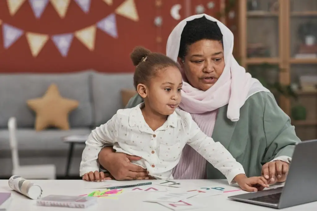 Muslim mother in hijab helping her young daughter with online learning at a laptop in their home.